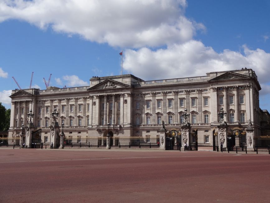 A large, historic stone building with classical architectural details, including columns and decorative balustrades, set against a backdrop of a partly cloudy blue sky. The building features multiple 