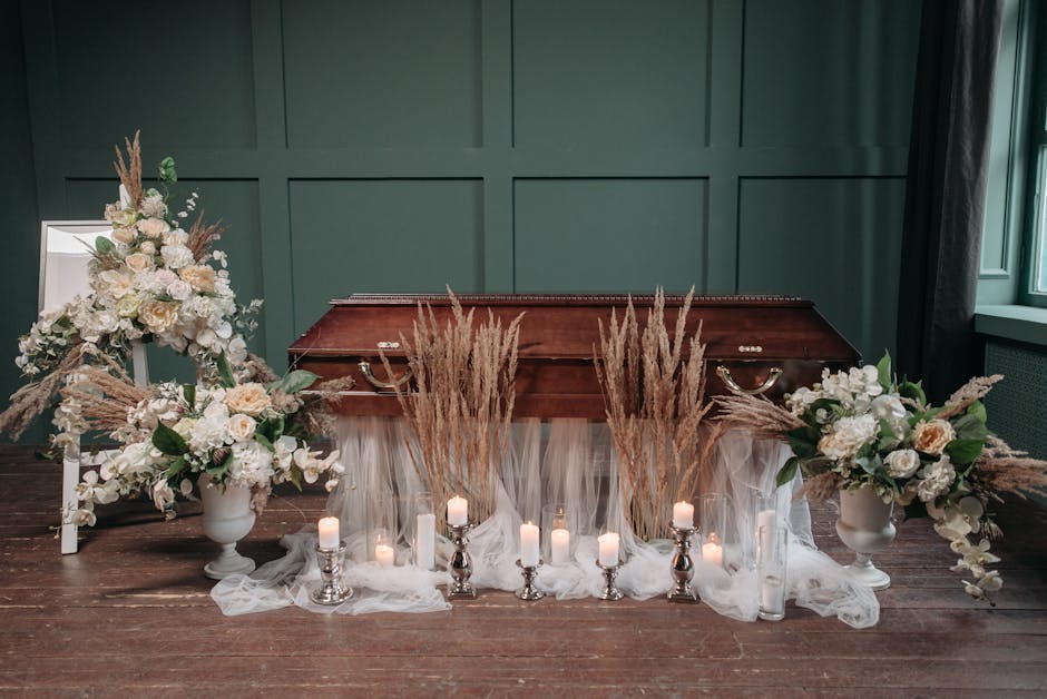 A floral arrangement for a funeral display featuring two large white urn vases filled with cream and white roses, hydrangeas, and eucalyptus, accented with tall beige pampas grass. The flowers are arr