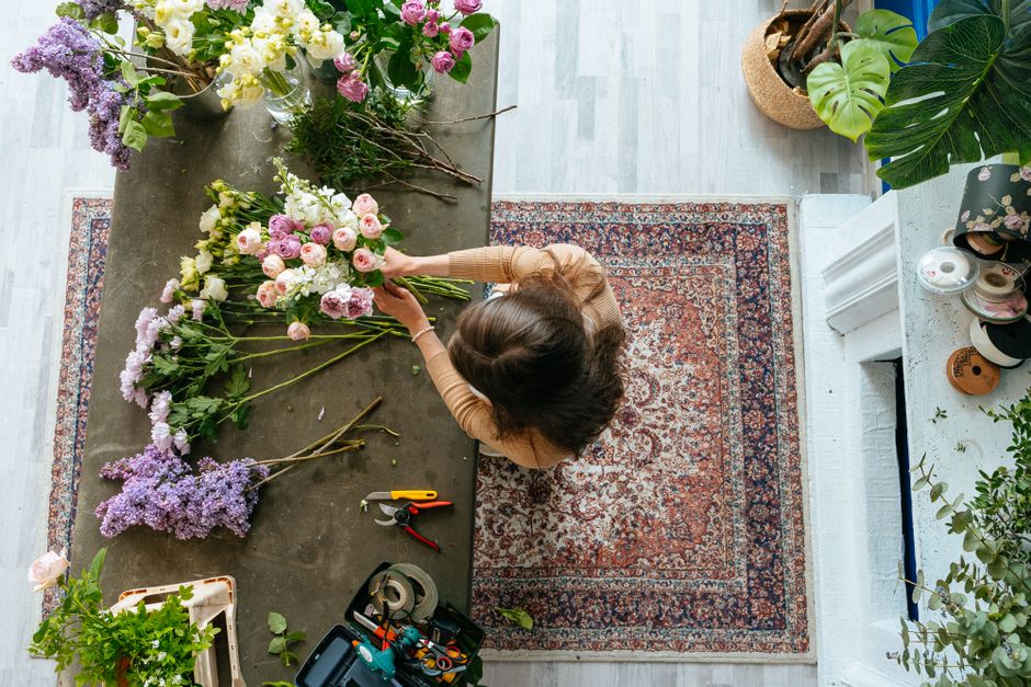 A florist with dark, wavy hair and wearing a beige long-sleeve top, seen from above, arranging a bouquet of fresh flowers on a dark grey work table. The bouquet includes pink lisianthus, white peonies