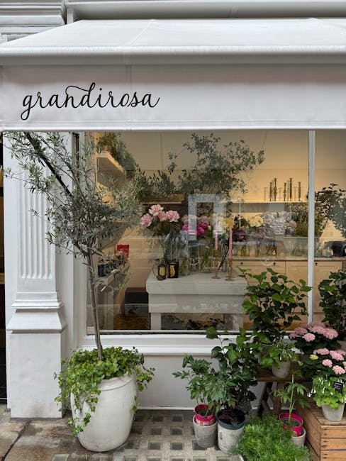 The image shows the exterior of a flower shop named 'Grandiroza' with a large glass window displaying floral arrangements inside. Outside the shop, there are several potted plants and flowers, includi
