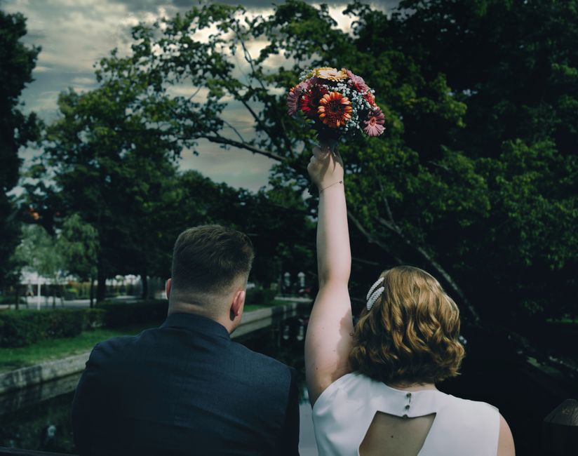 A woman with wavy blonde hair, wearing a white dress with a cut-out detail on the back, holds a colorful bouquet of fresh flowers high in the air during an outdoor event. The bouquet consists of vibra