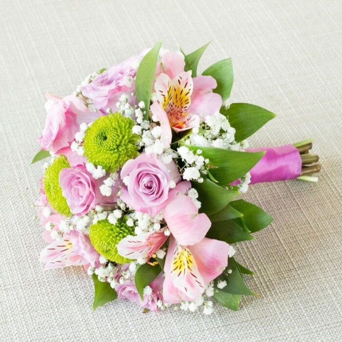 This exquisite hand-tied bouquet captures the refined essence of St James's, arranged diagonally atop a luxuriously soft, light beige fabric that recalls the classic textiles found in local boutiques. At its heart are pastel pink roses, each bloom inviting the senses with its delicate, spiralled petals and hint of subtle fragrance. Intertwined with the roses are bright green button chrysanthemums, whose compact, spherical forms inject a playful vibrancy and a modern twist to the palette. Slender stems of pale pink alstroemeria open to reveal intricate golden-yellow and burgundy-splashed petals, introducing a graceful, exotic note. Wisps of baby's breath lend the whole bouquet a feathery, ethereal quality, seamlessly linking each flower into a harmonious unity. Lush, dark green foliage frames the bouquet, giving a fresh, garden-picked look reminiscent of St James's local parks. The stems are neatly trimmed and elegantly wrapped in a deep magenta satin ribbon for a touch of stately glamour. Bathed in softly diffused daylight, every petal and leaf stands out with crisp freshness unique to central London floristry. Perfect for weddings or anniversaries celebrated near Piccadilly, this bouquet is an expression of romance, artistry, and the high standards that define St James's, crafted with a florist's devotion to every detail.