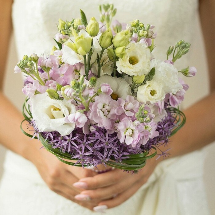 Captured in a gently focused composition, this image showcases a modern bridal bouquet as lovingly arranged by experienced florists in St James's. The camera frames the lower half of a bride's hands, nails carefully polished in delicate pink, cupping the bouquet just above its concealed stem wrap. Hints of a flowing white or lace-embroidered wedding gown shimmer in the background-a subtle nod to the timeless grandeur of venues like St James's Church or private West End salons. The bouquet itself is a lavish, rounded gathering of flower types and hues chosen to express both elegance and playfulness: large, ruffled white Lisianthus blooms with delicate greenish-yellow hearts dominate, while light pink and lilac stock blossoms infuse the design with soft texture and romantic pastel brilliance. Fresh green buds and slender spikes of foliage add vibrancy and depth, much like the floral borders found in the nearby royal park. Anchoring the arrangement, a wide swath of vivid purple, star-shaped Allium introduces a contemporary geometric twist, reminiscent of the district's mix of heritage and modernity. Elegantly curled bear grass-its deep green blades spiraling at the bouquet's base-lend sculptural interest, showcasing the floral artistry for which St James's florists are known. This bouquet, radiating warmth, sophistication, and fresh fragrance, is tailored for a couple celebrating among the historic avenues and joyful streets of central London.