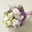 A sumptuous hand-tied bouquet reflecting the storied elegance of St James's, crafted with artistry and care on a lightly textured, neutral surface. At its core are generous white Lisianthus blooms, their creamy yellow centers and supple petals announcing a spirit of romance, surrounded by unopened buds that speak of hope and anticipation. Gentle waves of Stock flowers in pastel pink and lavender tones encircle the arrangement, creating a ruffled, frothy texture reminiscent of clouds floating over St James's Park on a spring morning. Striking purple Allium 'Star of Persia' emerge within the bouquet, their spiky globes adding texture and artistry that bridge tradition and modernity in the spirit of local Westminster galleries. The interplay of vivid green foliage-including slender leaves and budding Lisianthus-provides lively structure and a sense of fresh abundance. Elegantly gathered and wrapped in a bold fuchsia satin ribbon, accented with a delicate pearl strand, the stems are as beautiful as the bouquet's crown. Illuminated by gentle daylight, this exquisite display would set a stunning scene for a St James's wedding, a sophisticated celebration, or any event seeking romance and refined charm in SW1.